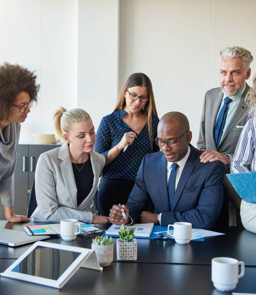 Group of focused businesspeople standing around their manager sitting at an office boardroom table reviewing charts and paperwork